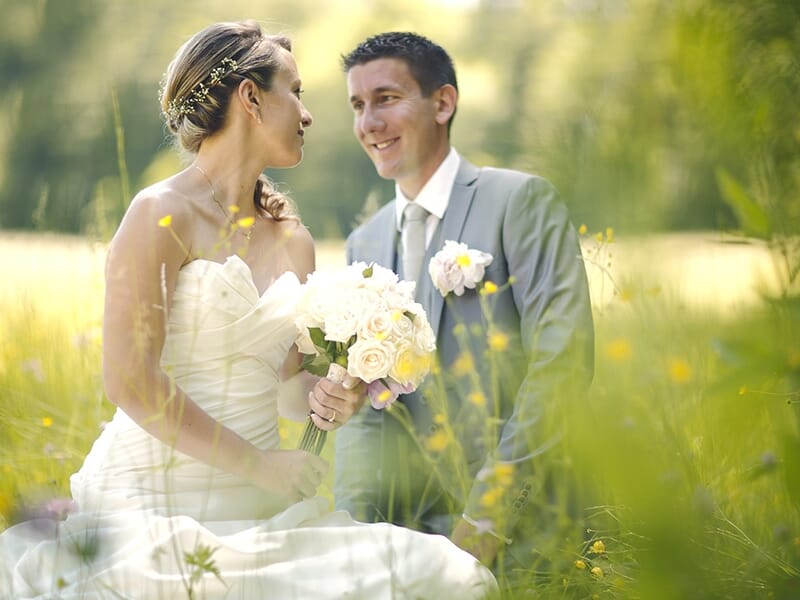 Mariés assis dans un champ fleuri, se regardant avec tendresse, la mariée tenant un bouquet de roses blanches.