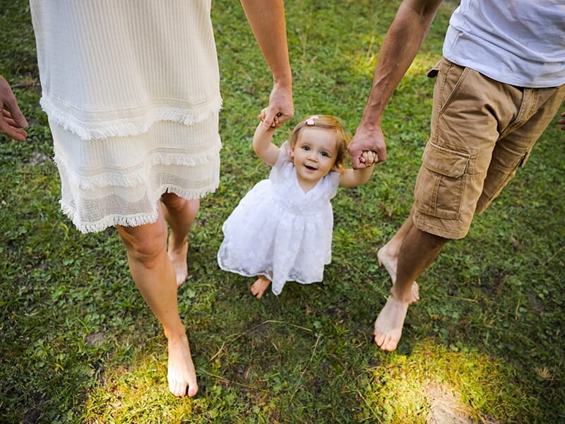 Petite fille en robe blanche marchant pieds nus dans l’herbe, tenant les mains de ses parents.