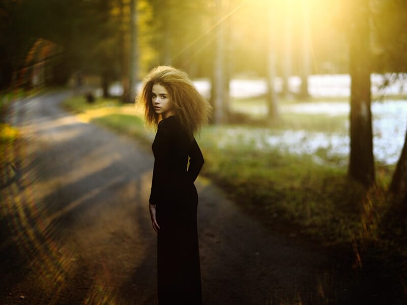 Jeune femme aux cheveux bouclés en robe noire, debout sur une route forestière baignée de lumière dorée.