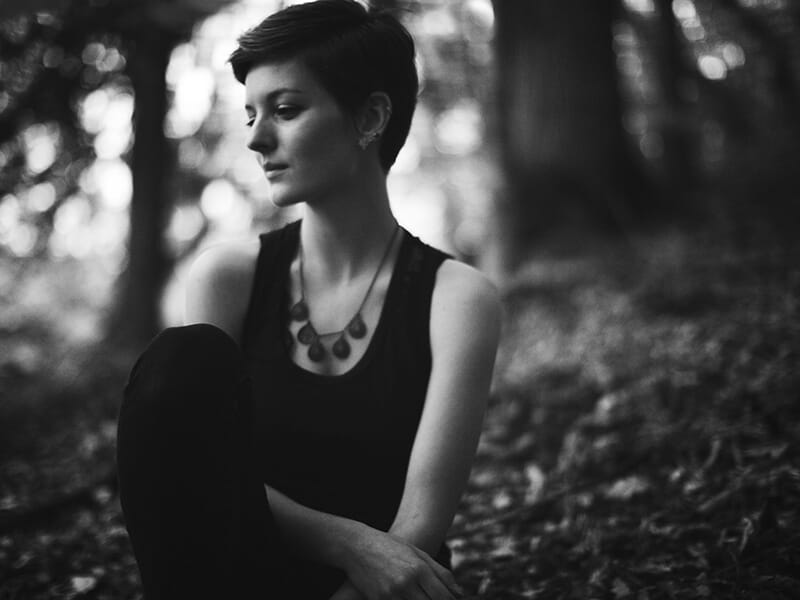Femme assise en forêt, en noir et blanc, les yeux baissés, dans une atmosphère calme et mélancolique.