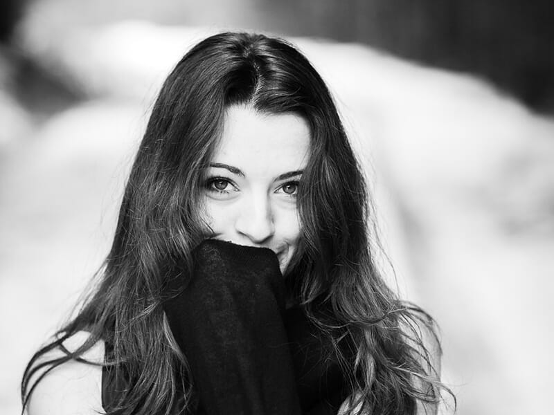 Jeune femme souriante cachant partiellement son visage derrière son pull, en noir et blanc, sur fond de chemin enneigé.