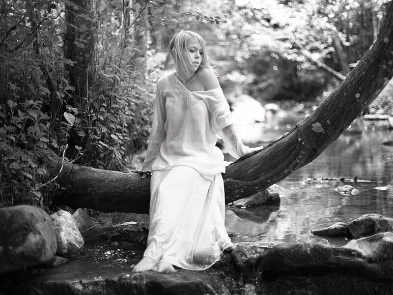 Femme en robe blanche assise sur un tronc d’arbre au-dessus d’un ruisseau, dans un décor boisé et paisible, en noir et blanc.
