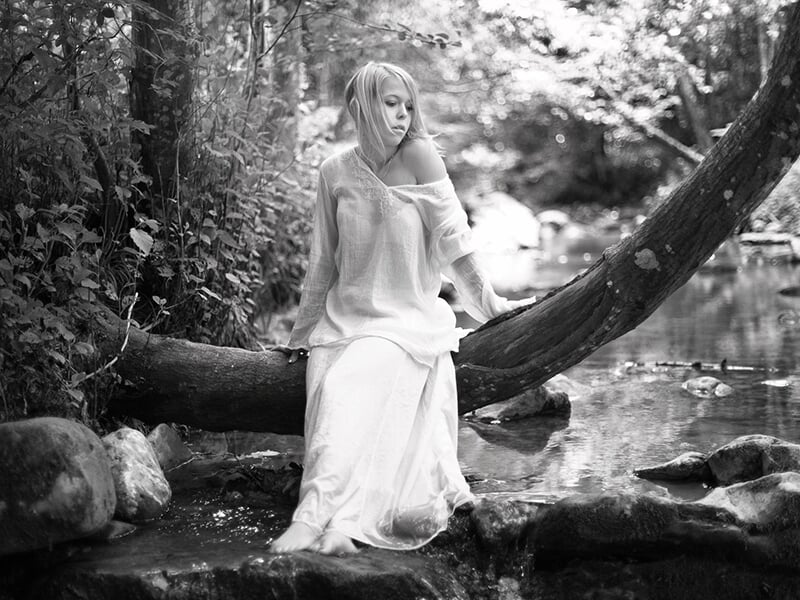 Femme en robe blanche assise sur un tronc d’arbre au-dessus d’un ruisseau, dans un décor boisé et paisible, en noir et blanc.