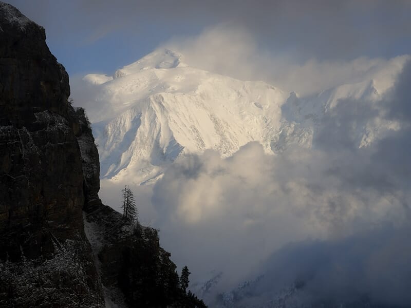 Sommet enneigé du Mont Blanc émergeant des nuages avec des parois rocheuses en premier plan.