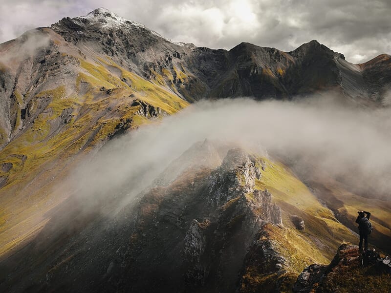Photographe en montagne devant une crête rocheuse surplombée de brume et de sommets aux teintes automnales.