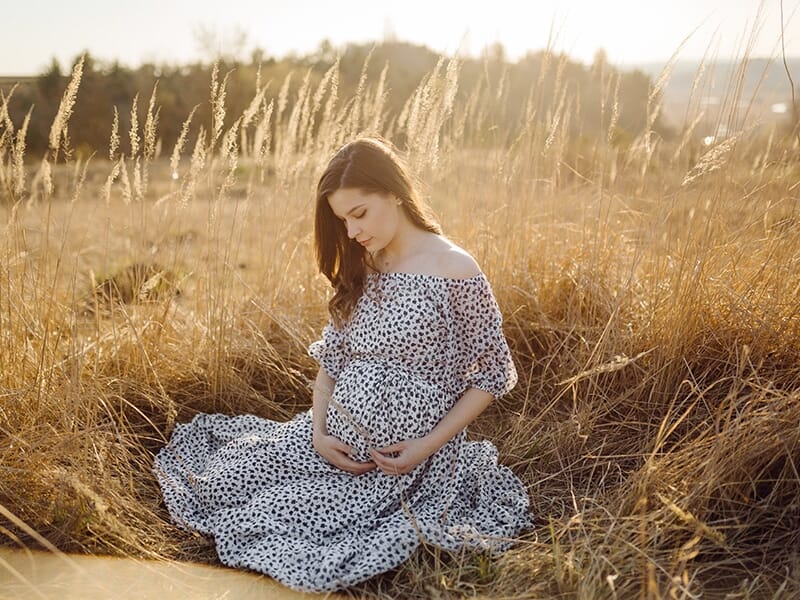 Portrait d'une femme enceinte assise dans un champ doré au coucher du soleil, les mains posées tendrement sur son ventre.