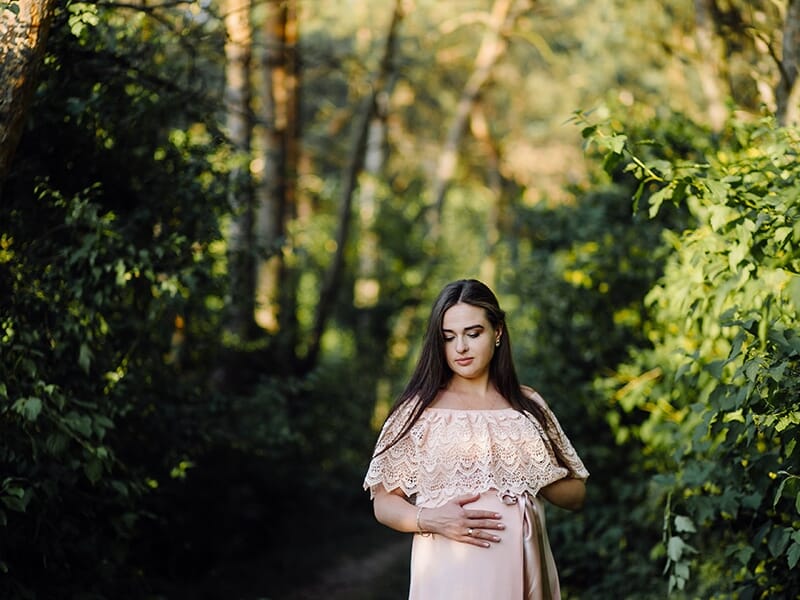 Femme enceinte en robe rose poudré, debout dans une forêt baignée de lumière, les mains posées sur son ventre arrondi.
