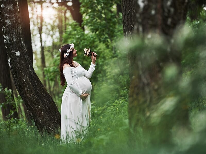 Femme enceinte en robe blanche et couronne de fleurs, debout dans une forêt verdoyante, tenant une fleur dans la main.