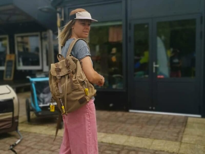 Femme portant un sac à dos marron avec cadenas jaunes, devant un bâtiment.