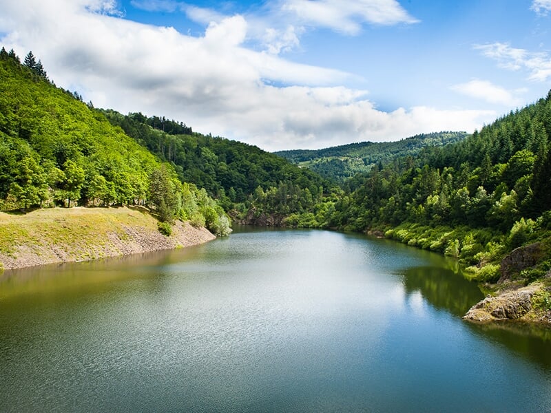 Vue panoramique d’un lac entouré de collines verdoyantes sous un ciel partiellement nuageux.