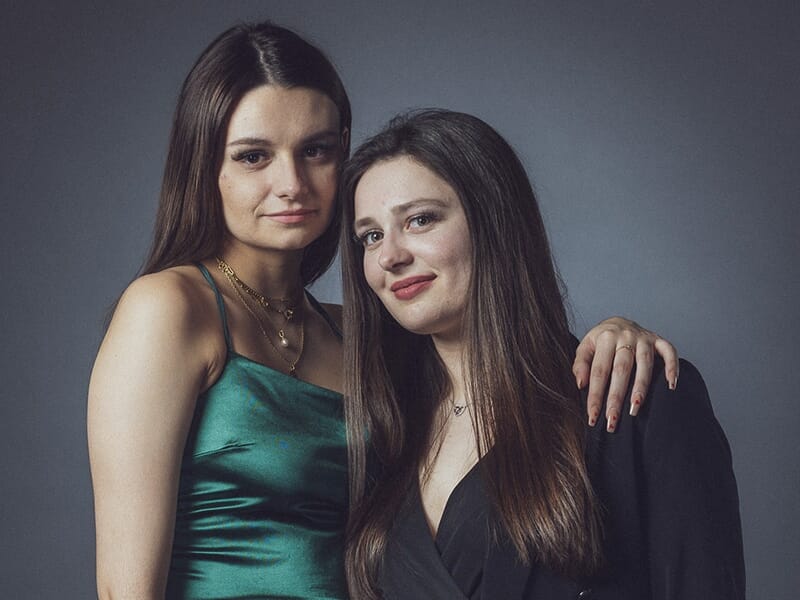 Deux jeunes femmes posent ensemble, l’une en robe verte satinée, l’autre en tenue noire, souriantes et complices.