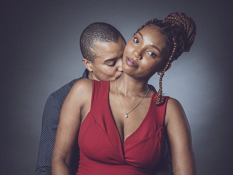 Un couple amoureux, la femme en robe rouge et l’homme l’enlaçant tendrement en l’embrassant dans le cou.
