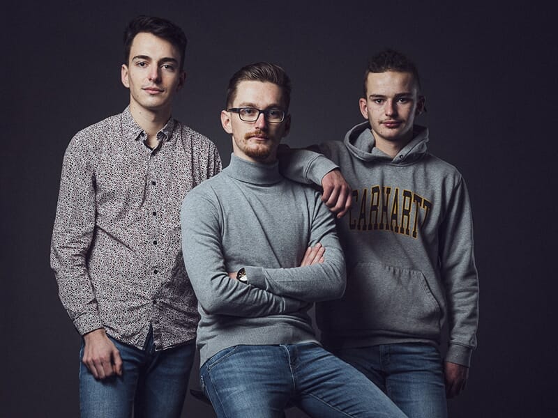 Trois jeunes hommes posent côte à côte, habillés décontractés, dans une ambiance sobre et amicale.