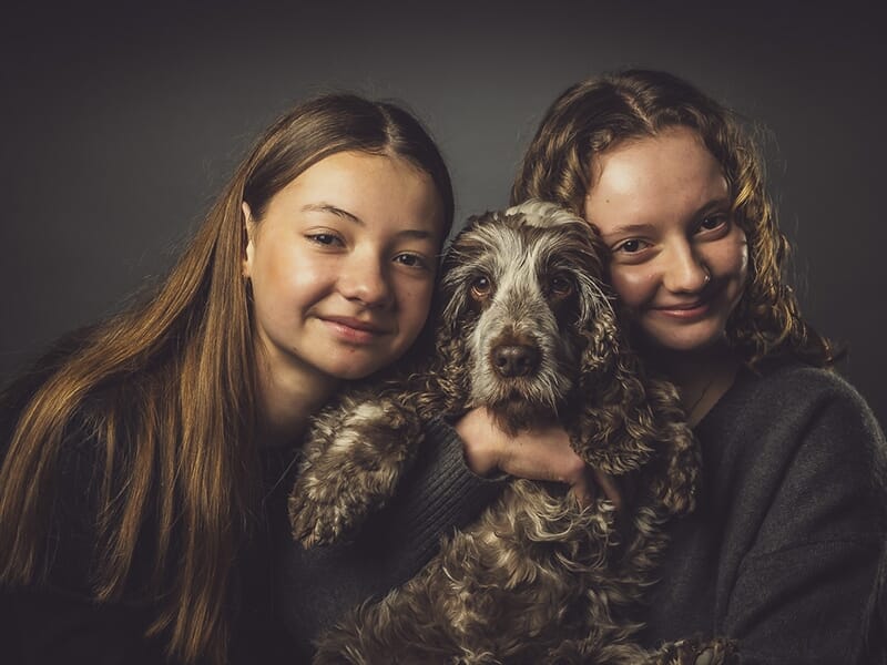 Deux jeunes filles serrent un chien dans leurs bras, tous les trois regardant l’objectif avec douceur.