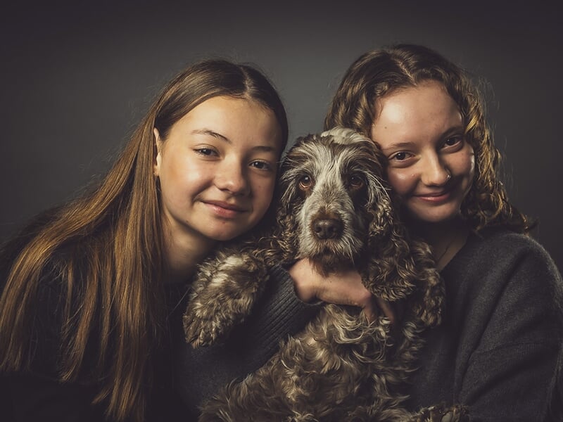 Deux jeunes filles serrent un chien dans leurs bras, tous les trois regardant l’objectif avec douceur.