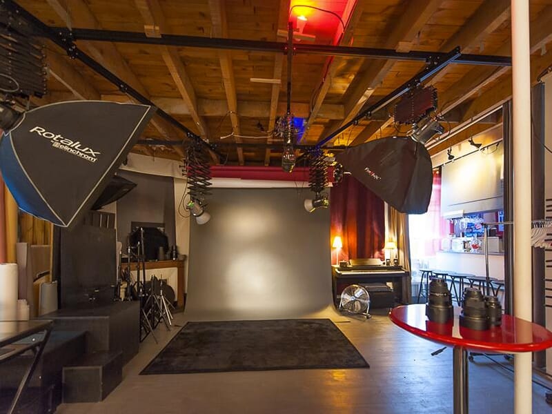 Espace de prise de vue professionnel avec fond gris, éclairages suspendus, réflecteurs et objectifs photo posés sur une table.