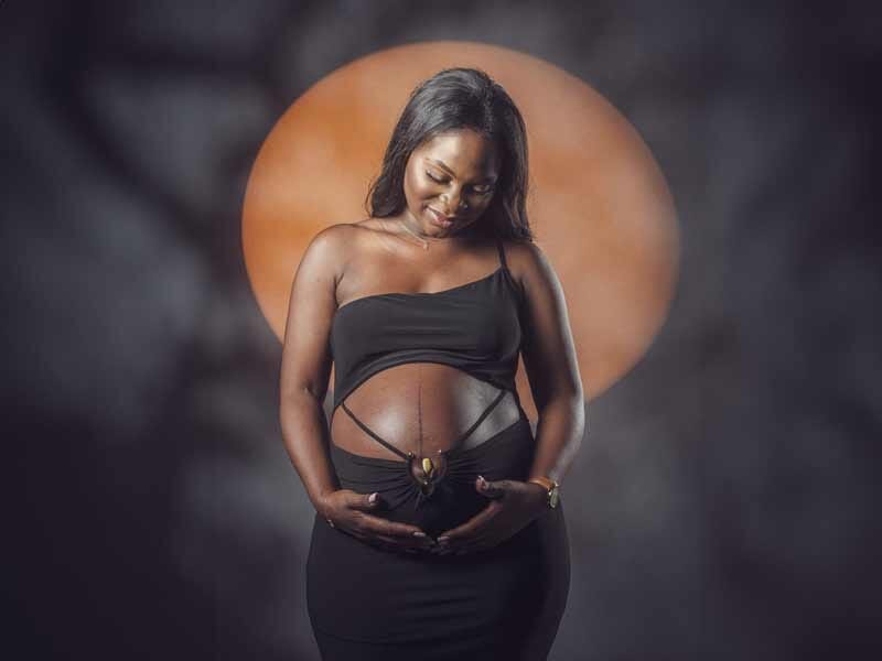 Femme enceinte souriante en robe noire tenant son ventre, avec un cercle orange lumineux en arrière-plan.