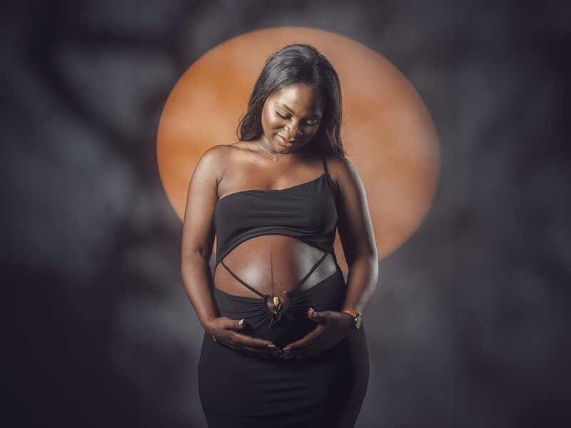 Femme enceinte souriante en robe noire tenant son ventre, avec un cercle orange lumineux en arrière-plan.