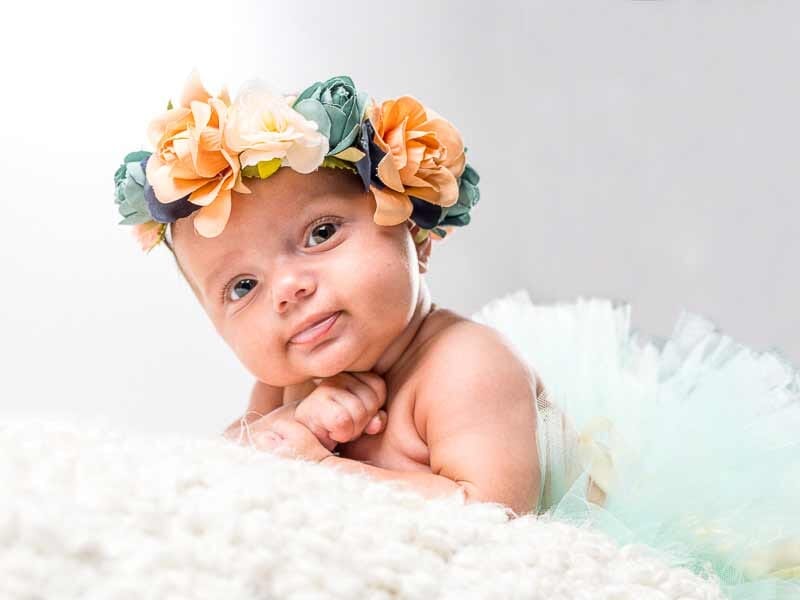 Bébé allongé sur un tapis blanc, portant une couronne de fleurs colorées, regard doux vers l'objectif.