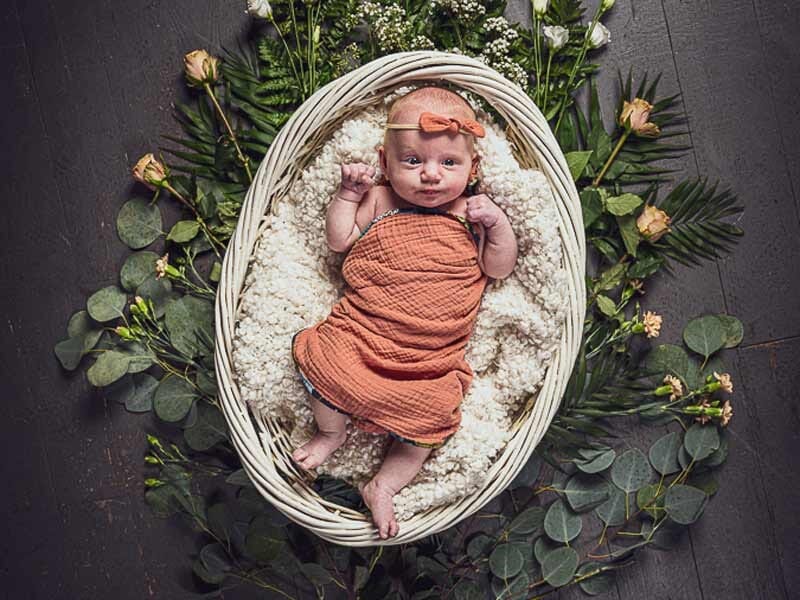 Nouveau-né emmailloté en orange dans un panier blanc, entouré de feuillages et de fleurs sur un sol sombre.