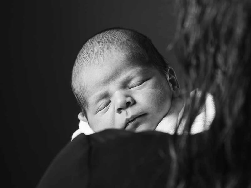 Portrait en noir et blanc d'un bébé endormi, posé sur l'épaule d'un adulte, ambiance douce et intime.