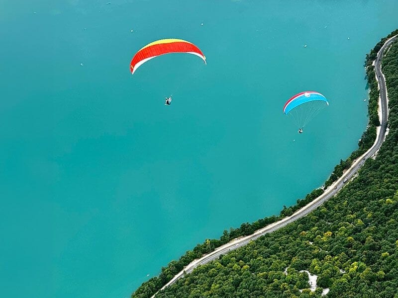 Deux parapentes colorés volent au-dessus du lac turquoise d'Aiguebelette avec une route et forêt en bordure.