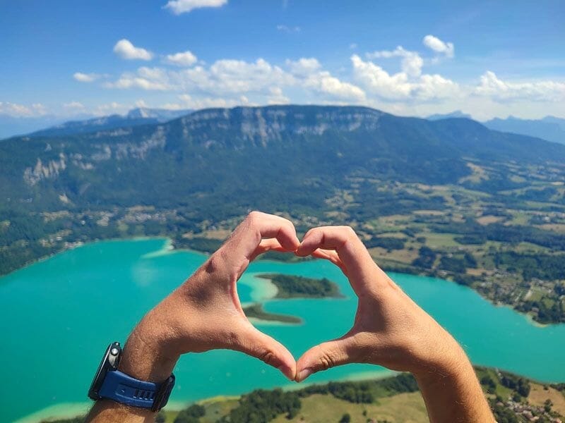Mains formant un cœur devant le lac d'Aiguebelette vu en hauteur avec montagnes et ciel bleu en arrière-plan.