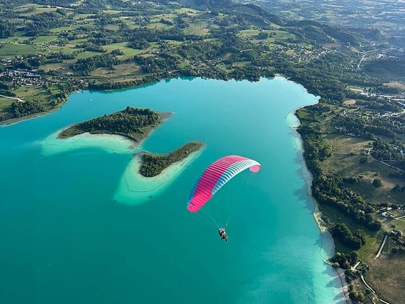 Parapente rose survolant le lac d'Aiguebelette avec îles et paysage naturel environnant sous un ciel clair.