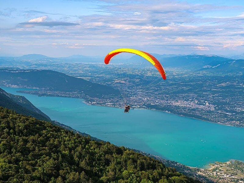 Parapente orange volant au-dessus du lac d'Aiguebelette avec vue sur la ville et montagnes au loin sous un ciel bleu.