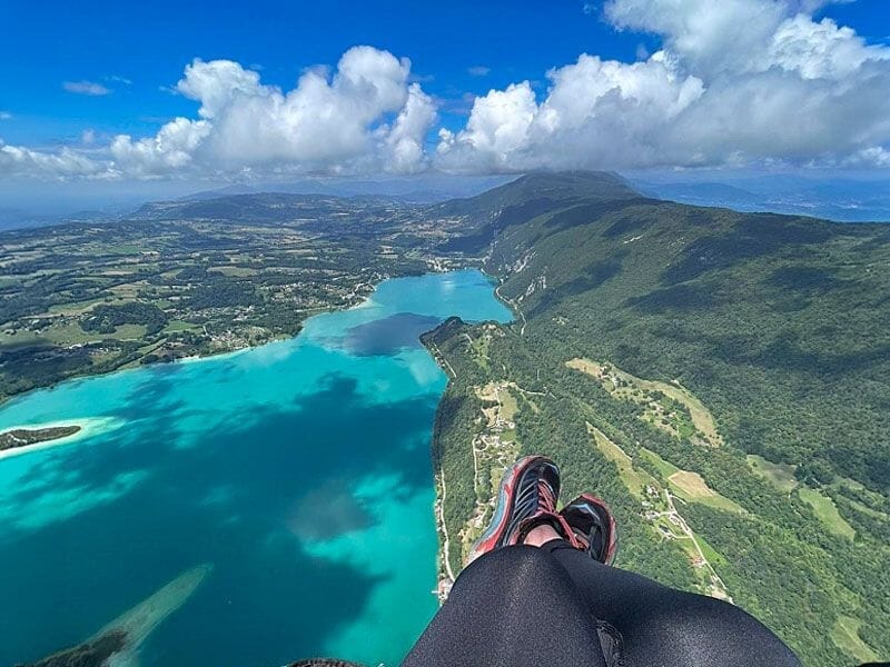 Vue en plongée sur le lac d'Aiguebelette et montagnes avec jambes d'un parapentiste en premier plan en vol.