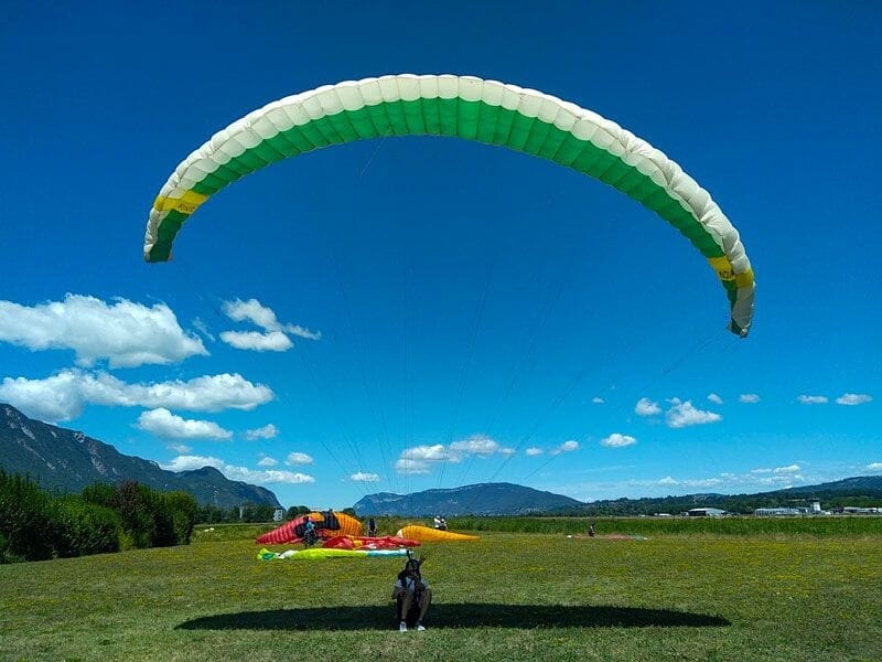 Parapentiste au sol avec voile verte et blanche déployée sur un champ fleuri entouré de montagnes et ciel bleu.