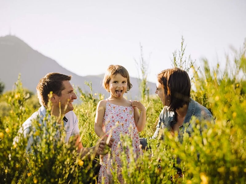 Petite fille souriante tirant la langue entre ses deux parents assis dans un champ ensoleillé, avec des montagnes en fond.
