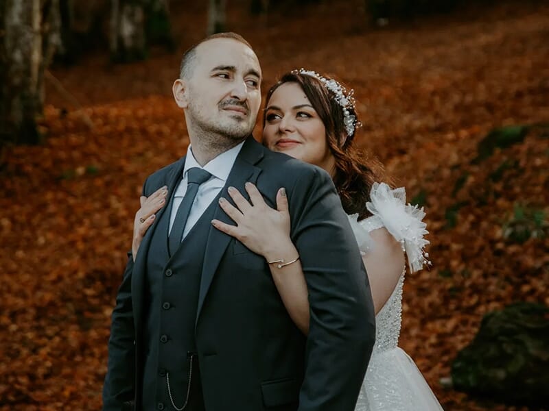 Couple de mariés enlacés dans une forêt d’automne, la mariée souriante tenant son époux par derrière.
