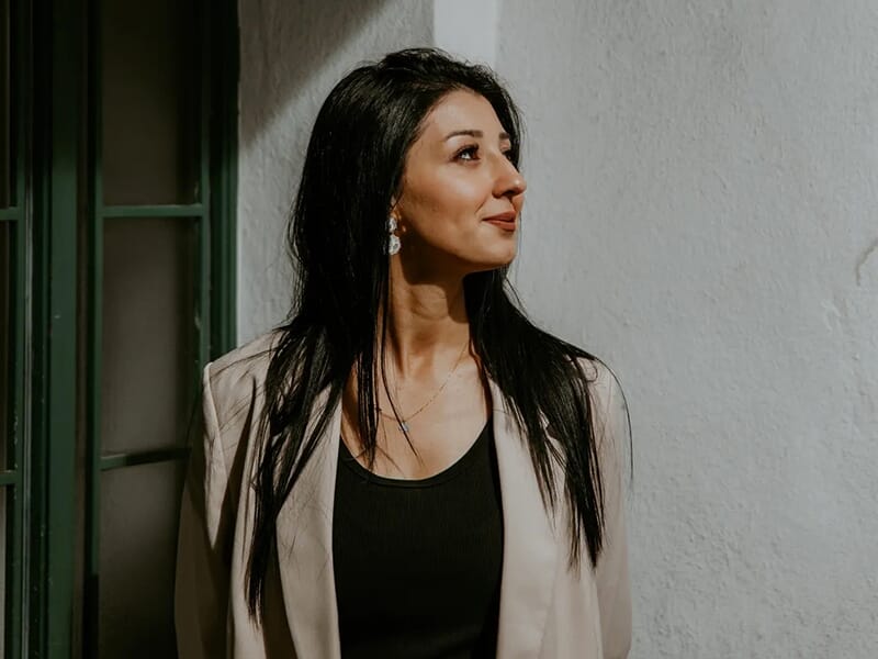 Femme aux cheveux noirs longs regardant sur le côté, debout près d’un mur blanc à la lumière douce.