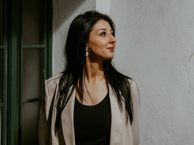  Femme aux cheveux noirs longs regardant sur le côté, debout près d’un mur blanc à la lumière douce.
