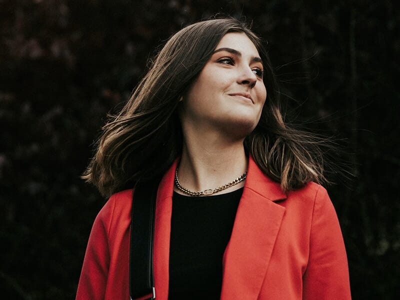 Jeune femme souriante en veste rouge et haut noir, posant en extérieur avec les cheveux au vent.