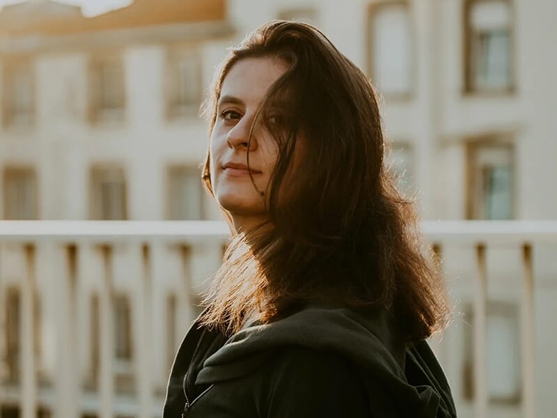 Personne aux cheveux mi-longs regardant par-dessus son épaule, en lumière dorée devant un bâtiment flou.