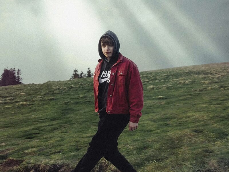Jeune homme en veste rouge et sweat à capuche marchant dans un paysage verdoyant sous un ciel dramatique.