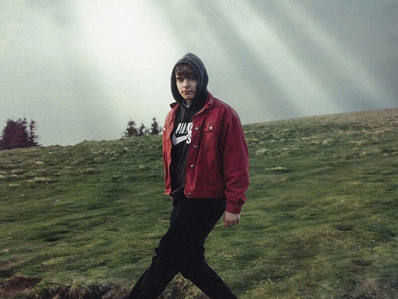 Jeune homme en veste rouge et sweat à capuche marchant dans un paysage verdoyant sous un ciel dramatique.