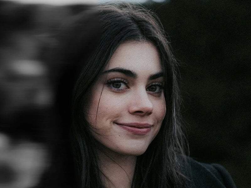Portrait serré d’une jeune femme au regard doux et aux cheveux noirs, sur fond sombre et flou.