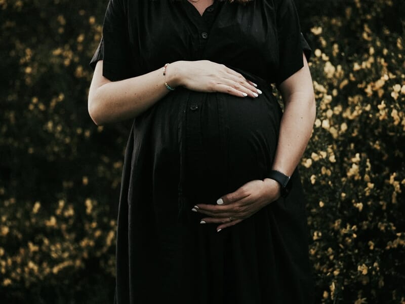 Femme enceinte en robe noire tenant son ventre avec tendresse, entourée de fleurs jaunes.