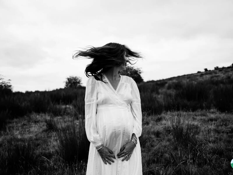  Femme enceinte en robe blanche regardant sur le côté, les cheveux au vent, dans un champ sauvage.