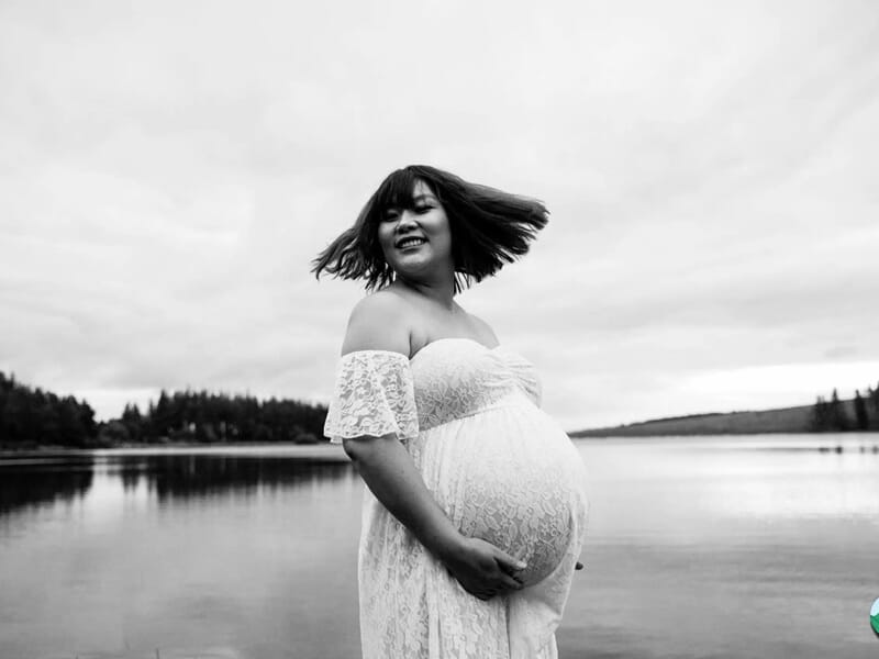 Femme enceinte souriante en robe blanche en dentelle, posant près d’un lac avec les cheveux en mouvement.