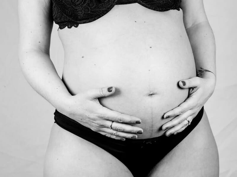 Portrait en noir et blanc d’une femme enceinte en lingerie noire, tenant son ventre à deux mains.