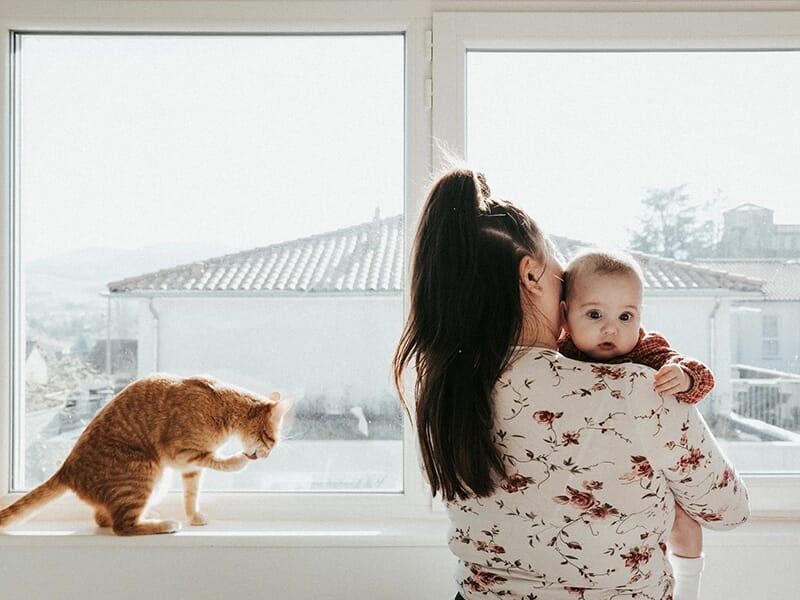 Maman portant son bébé face à une grande fenêtre, avec un chat roux posé sur le rebord.