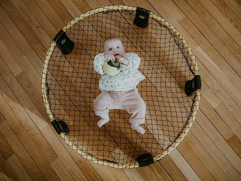 Bébé allongé au centre d’un filet circulaire sur un sol en bois, tenant un jouet dans ses mains.