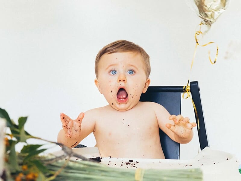 Bébé torse nu avec une expression de surprise, assis devant un gâteau d’anniversaire et des décorations festives.