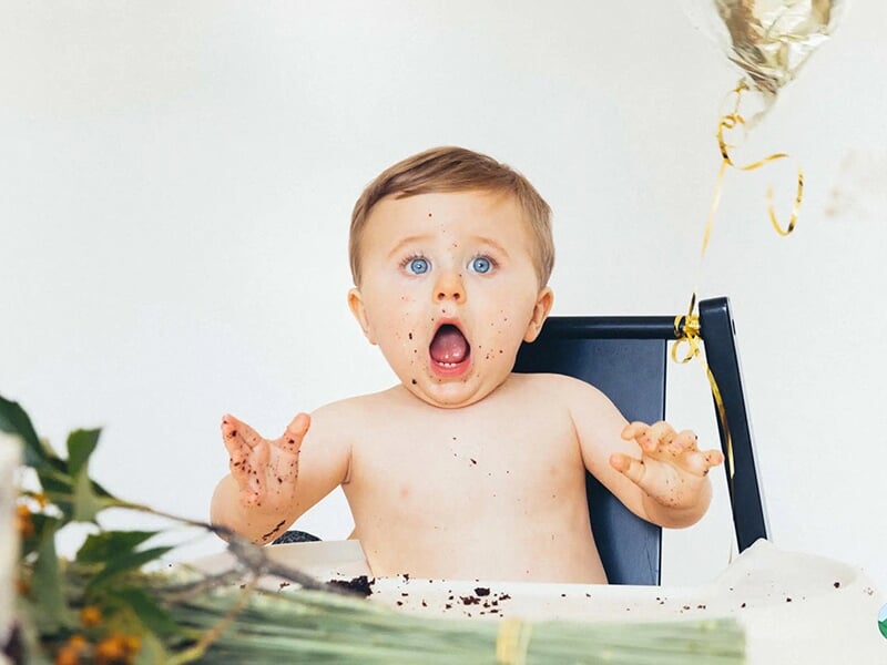Bébé torse nu avec une expression de surprise, assis devant un gâteau d’anniversaire et des décorations festives.