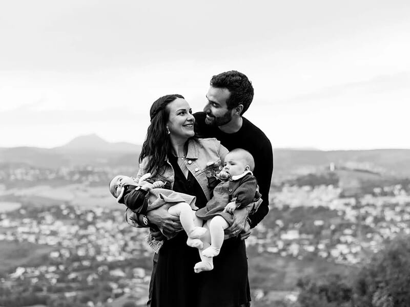 Couple souriant portant deux nourrissons dans les bras, avec une vue panoramique d’une ville en arrière-plan (photo en noir et blanc).