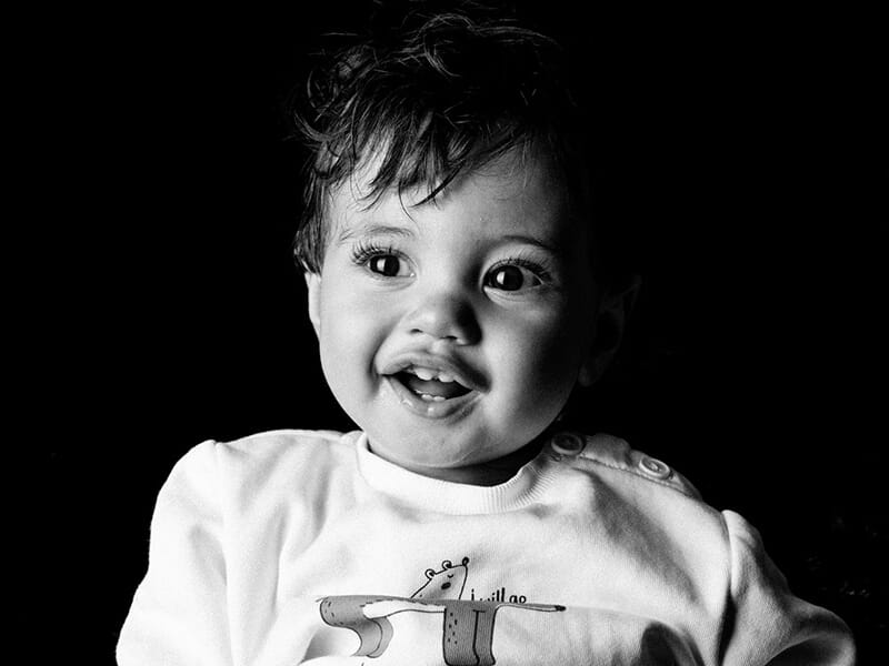 Portrait en noir et blanc d’un bébé souriant, capturé en gros plan sur fond sombre.